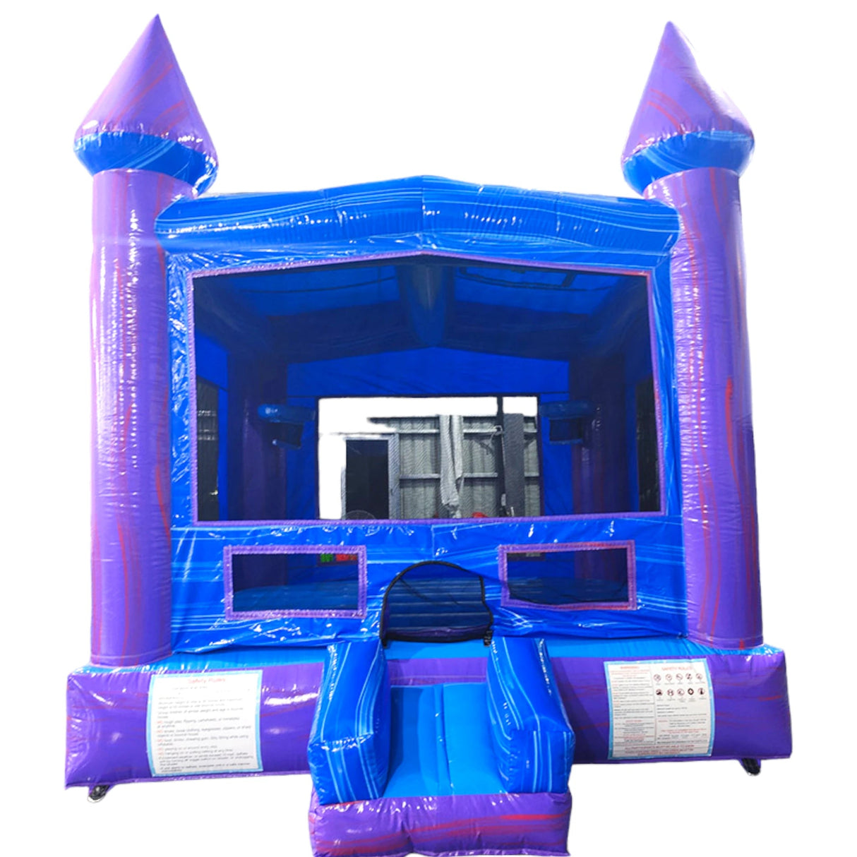 Front view of Mystic Commercial Grade Bounce House featuring a vibrant blue and purple color scheme. The inflatable structure has a castle-like design with two tall purple turrets topped with blue conical roofs and yellow flags. The main bounce area has blue walls with mesh windows for visibility. A blue curved slide extends from the right side. The entrance has a small step and an arched doorway.