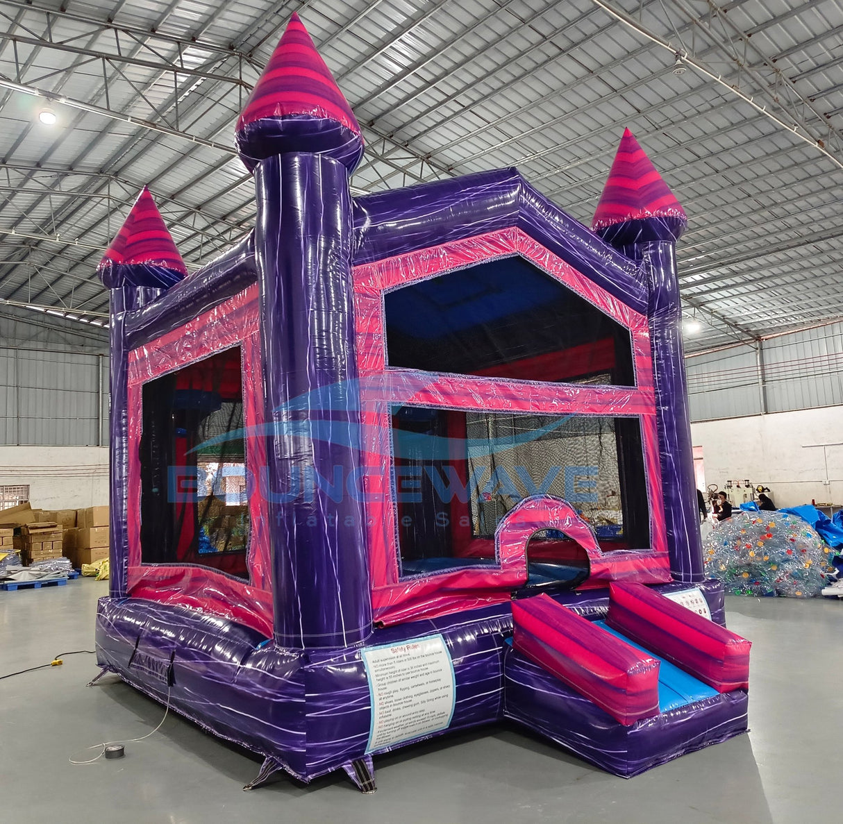 Side angle view of Purple Palace Commercial Grade Bounce House set up in a large indoor warehouse. The inflatable features purple walls, pink accents, and three tall towers with pink conical tops. Mesh windows allow visibility into the bounce area. The warehouse setting shows metal roofing, fluorescent lighting, and other inflatable products in the background.