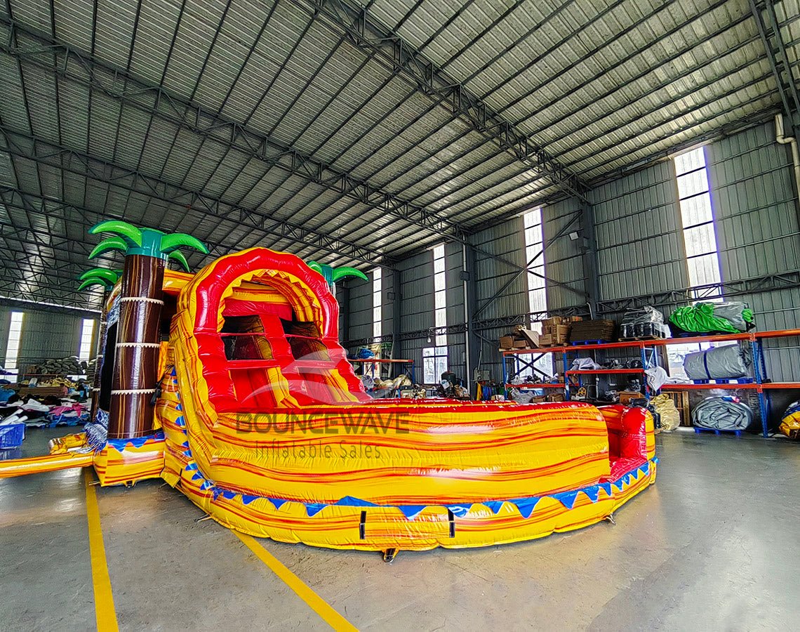 Angled front view of Rasta Curved Dual Lane Commercial Inflatable Combo (Wet/Dry) in a warehouse. Highlights the vibrant red and orange dual slides, blue castle entrance with palm trees, and expansive splash pool. Warehouse interior visible with metal roofing, shelving units, and other equipment, providing scale and context for the inflatable's size.