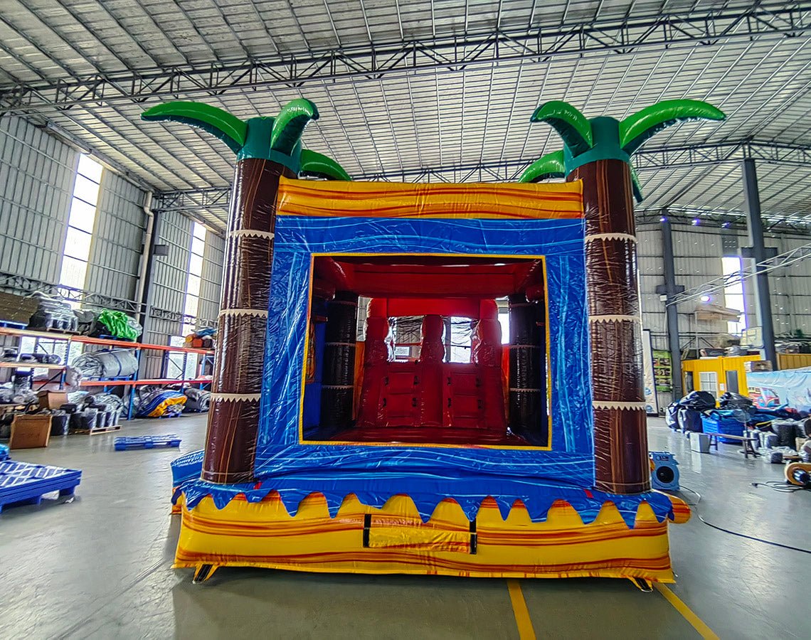 Wide-angle view of Rasta Curved Dual Lane Commercial Inflatable Combo (Wet/Dry) in a large indoor warehouse. Features a vibrant tropical-themed design with dual curved slides in red and orange, leading to a spacious red splash pool. The main structure has a blue castle-like entrance with palm tree decorations and yellow accents. Inflatable palm trees top the structure. The warehouse setting shows metal roofing, hanging disco balls, and other inflatable products in the background, providing scale and context