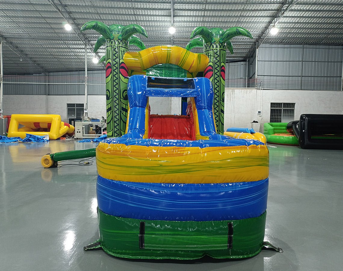 Front view of Reggae Rush Commercial Econo Inflatable Combo (Wet/Dry) highlighting the entrance with tiki face designs and palm tree decorations. The inflatable features a yellow slide and blue splash pool area. Set up on a polished concrete floor in an indoor warehouse, showcasing its vibrant tropical and rasta-inspired color scheme.