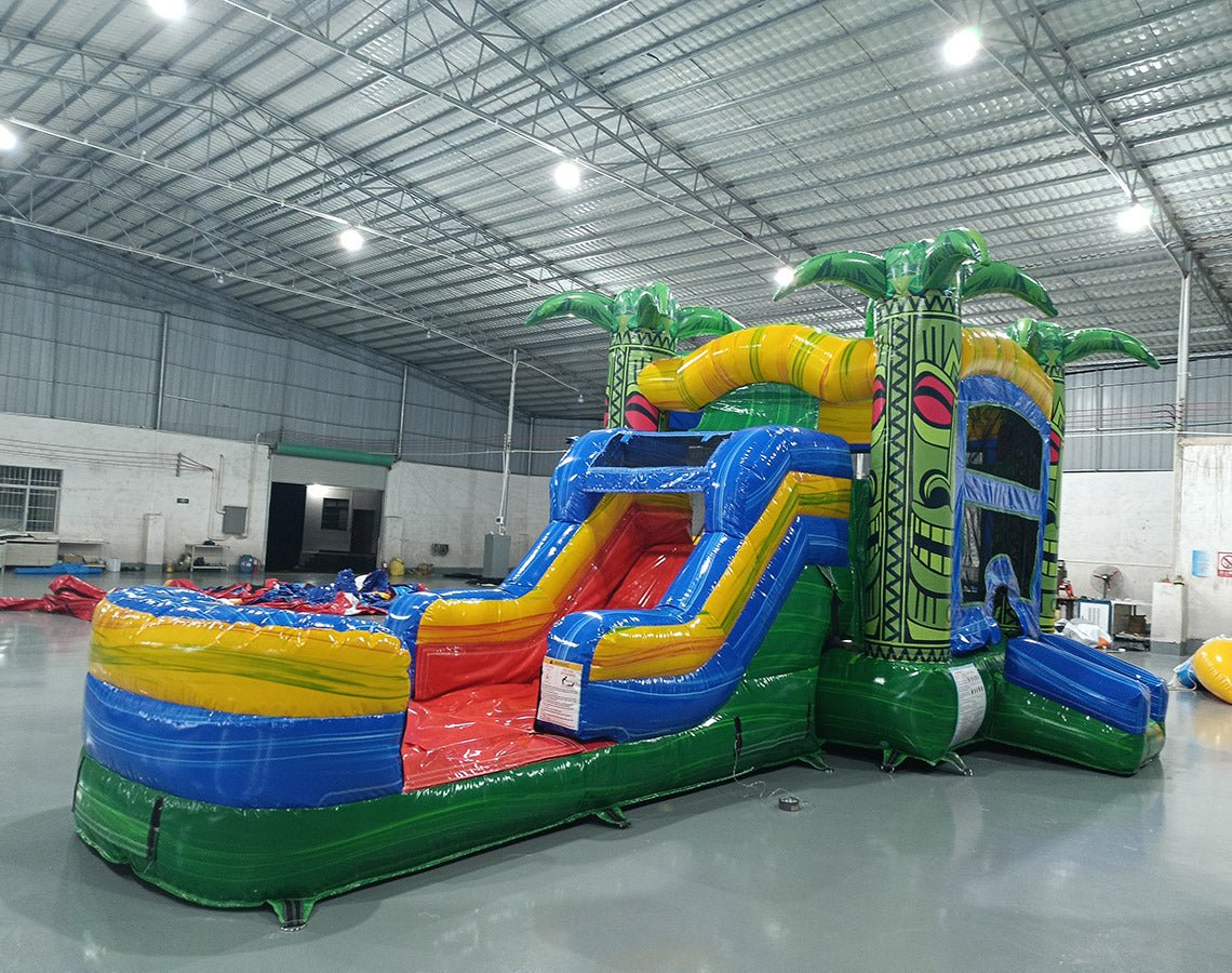Side view of Reggae Rush Commercial Econo Inflatable Combo (Wet/Dry) set up in a large indoor warehouse. Features tiki face designs, palm tree decorations, yellow slide, and blue splash pool area. The inflatable showcases vibrant green, yellow, blue, and red colors. Metal roofing, fluorescent lighting, and other inflatable products visible in the background.