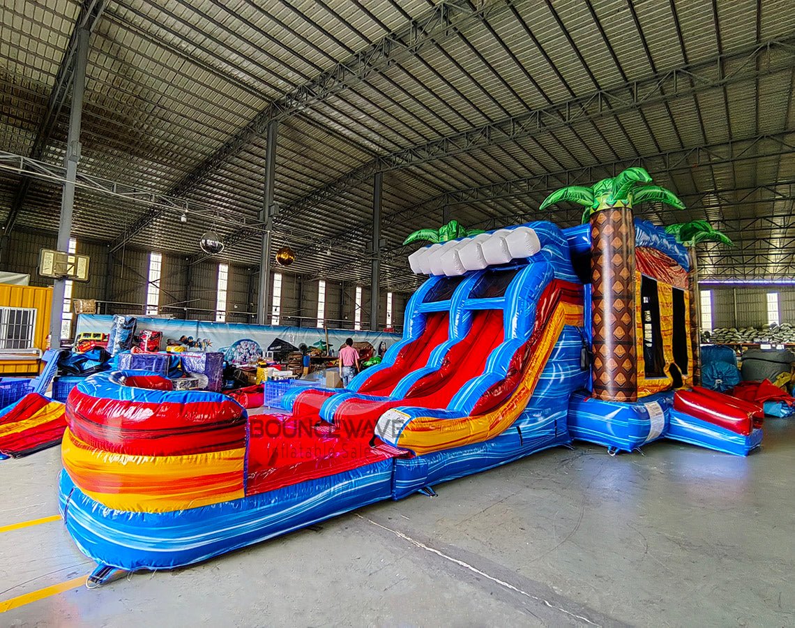 Wide-angle view of Rip Curl Dual Lane Commercial Inflatable Combo (Wet/Dry) set up in a large indoor warehouse. The inflatable displays dual blue slides, brown palm tree trunks with blue leaves, a yellow and blue bounce house area, and a large circular splash pool with rainbow-colored rings. Various other inflatable products, storage containers, and warehouse equipment are visible, showcasing its size and commercial setting.