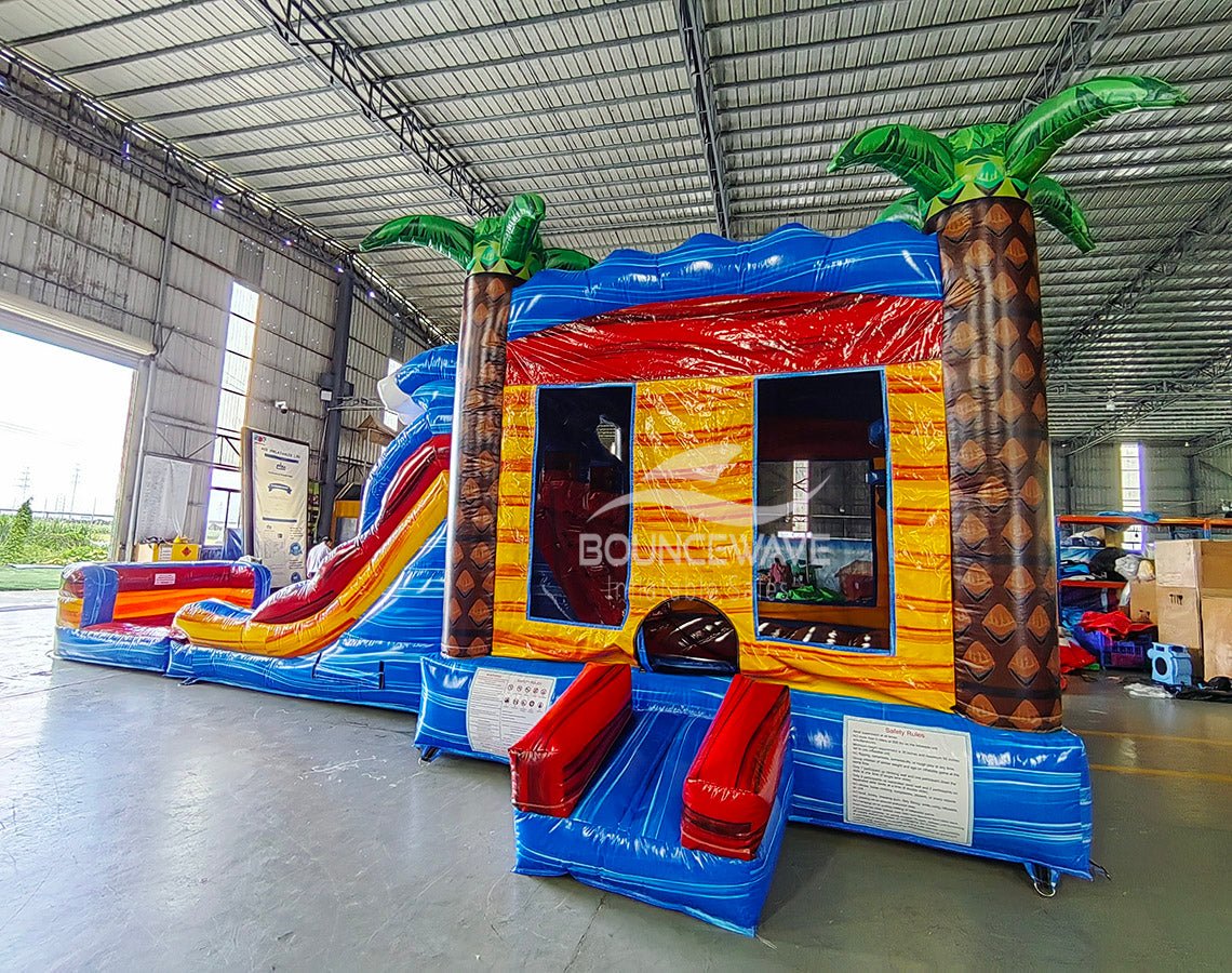Side view of Rip Curl Dual Lane Commercial Inflatable Combo (Wet/Dry) showing its full length. The inflatable features brown palm tree trunks with blue leaves, dual blue slides, a yellow and blue bounce house structure, and a large circular splash pool with rainbow-colored rings. Set in a large indoor warehouse with metal roofing, concrete flooring, and various other inflatable products visible in the background.