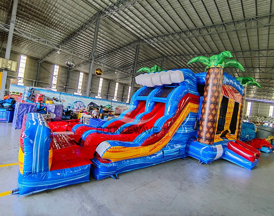 Wide-angle view of Rip Curl Dual Lane Commercial Inflatable Combo (Wet/Dry) in a spacious warehouse. The inflatable displays brown palm tree trunks with green leaves, dual blue slides, a bounce house area, and a large circular splash pool with vibrant rainbow-colored rings. Various other inflatable products, storage containers, and warehouse equipment are visible, showcasing its size and commercial setting.