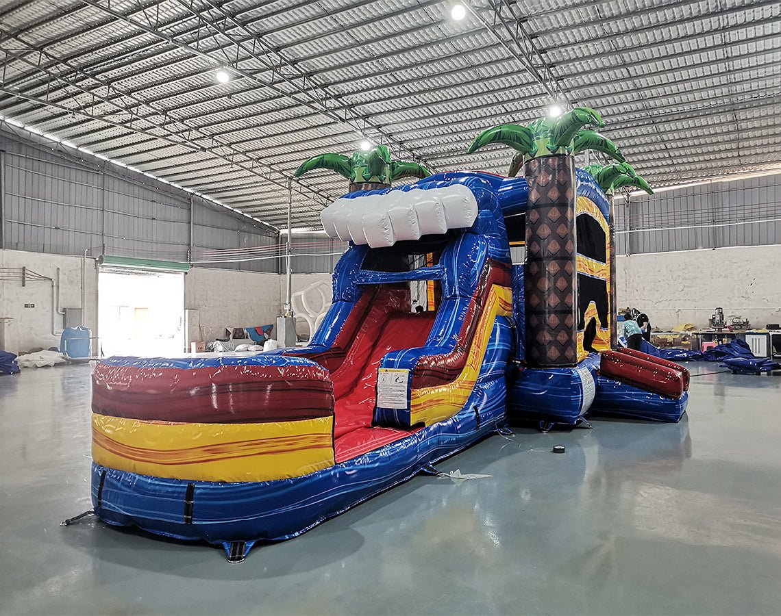 Side view of Rip Curl Econo Combo showing bright blue, yellow, and red design with inflatable palm trees, curved slide, and large splash pool area. The combo includes a bounce house and is set up in an indoor warehouse with metal roofing and other equipment visible.