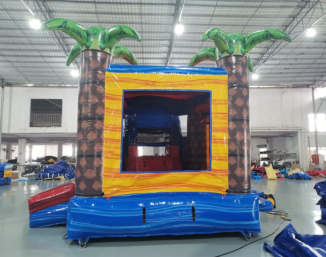 Large inflatable tropical-themed combo featuring a bounce house with palm tree decorations, attached slide, and splash pool. The structure has vibrant blue, yellow, and red colors with brown palm tree trunks. Set up in an indoor warehouse with metal roofing visible.
