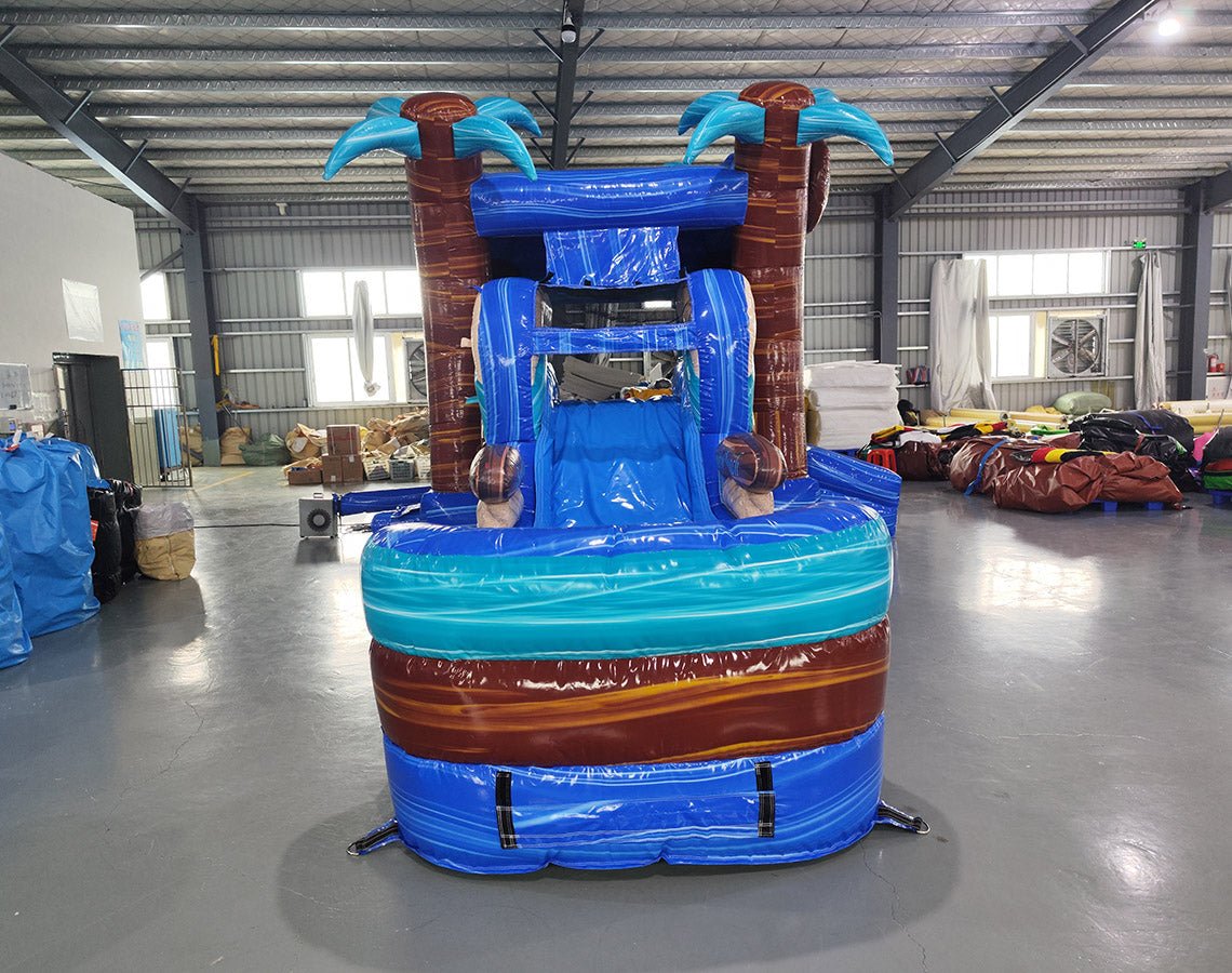 Large inflatable tropical-themed combo featuring a bounce house with palm tree decorations, attached slide, and splash pool. The structure has vibrant blue and turquoise colors with brown palm tree trunks and a large inflatable sunglasses decoration on top. Set up in an indoor warehouse with metal roofing visible.
