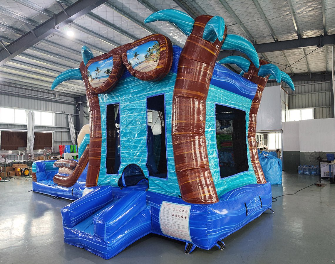Angled view of the South Beach Econo Combo featuring a blue and turquoise bounce house with palm tree decorations, slide, and spacious splash pool. The inflatable includes a large sunglasses decoration on top and is set up in an indoor warehouse with other equipment and inflatables visible.