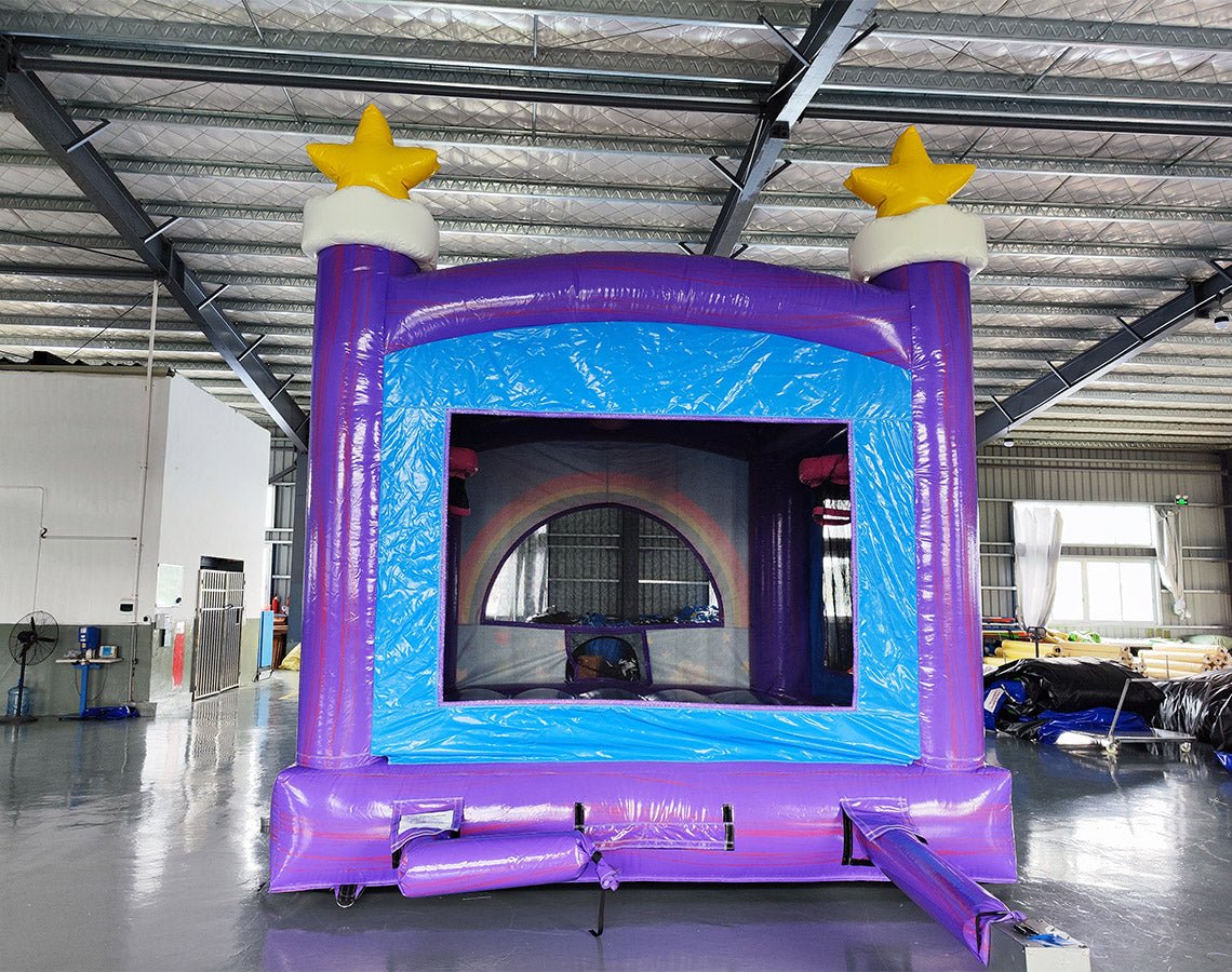 Rear view of Unicorn Commercial Grade Bounce House in a large indoor warehouse. The inflatable features a vibrant purple base with blue sky-patterned panels. A large white unicorn figure with a colorful mane sits atop a rainbow arch entrance. Two purple towers with yellow stars flank the sides. A small blue slide protrudes from the entrance. The warehouse setting shows metal roofing, windows, and various equipment and materials in the background.