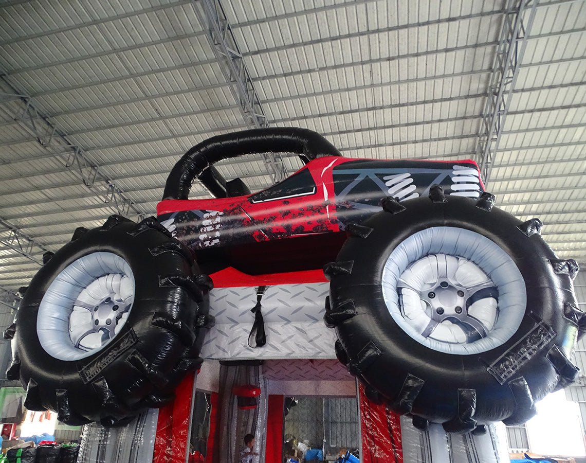 Side angle view of UTV Trail Boss Commercial Grade Bounce House in a warehouse setting. The inflatable off-road vehicle design shows detailed side panels, large black tires, and a roll cage. The red bounce house entrance is visible beneath the vehicle. Metal roofing, windows, and other inflatable products can be seen in the background.