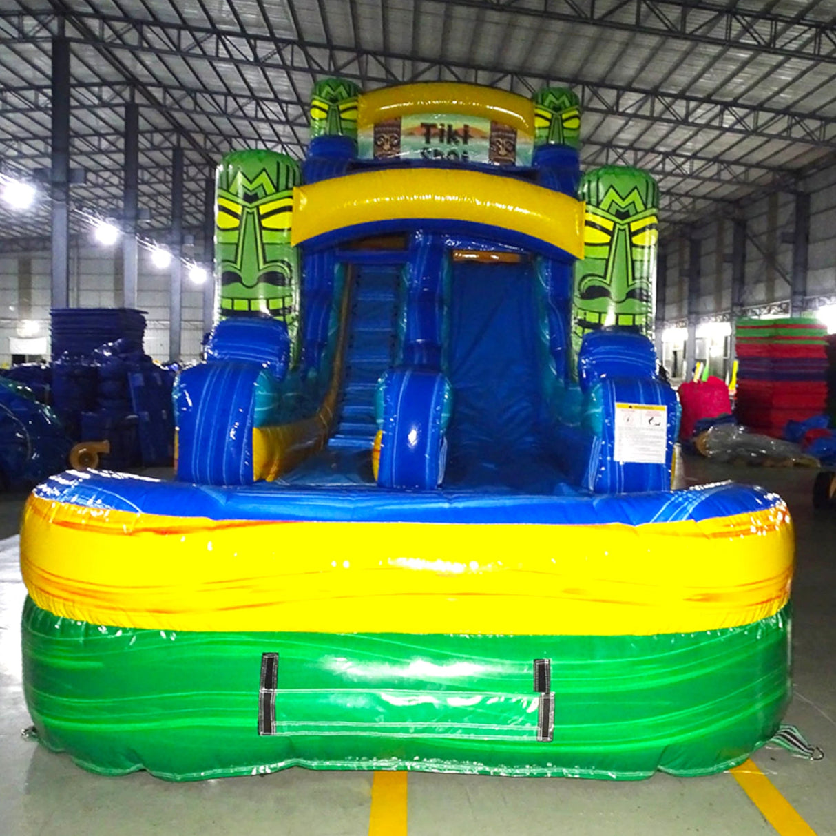 Front view of Tiki Shot single lane water slide in indoor warehouse, showing tall structure with blue sliding surface, tiki mask designs, and large splash area with yellow and green colors.