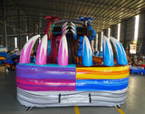 Front view of the 18ft Dino Duel inflatable water slide, highlighting its dual lanes in pink and blue, central climbing structure, dinosaur decorations, and inflatable palm trees, displayed in a large indoor space with other inflatables visible.
