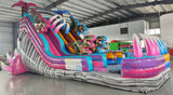 Full-length view of the 18ft Dino Duel Center Climb water slide, showcasing its colorful design with pink and blue lanes, dinosaur figures, palm trees, and a large splash pool area at the base, set up in an indoor facility.