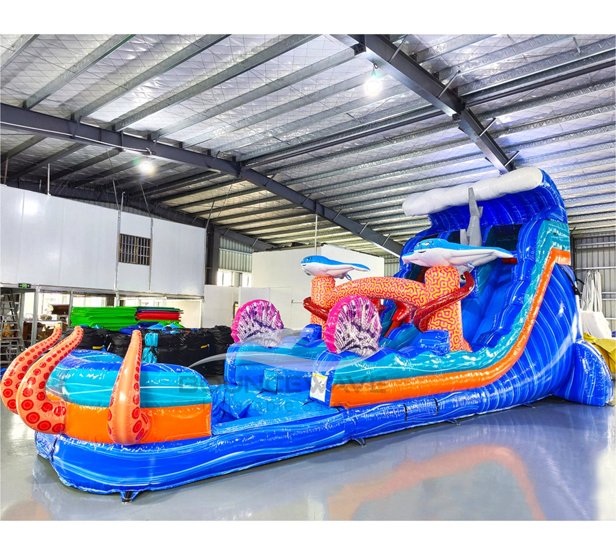Side view of Hammerhead Reef hybrid water slide in warehouse, showcasing its length, dual blue lanes, shark decorations, and large splash area with octopus tentacles.
