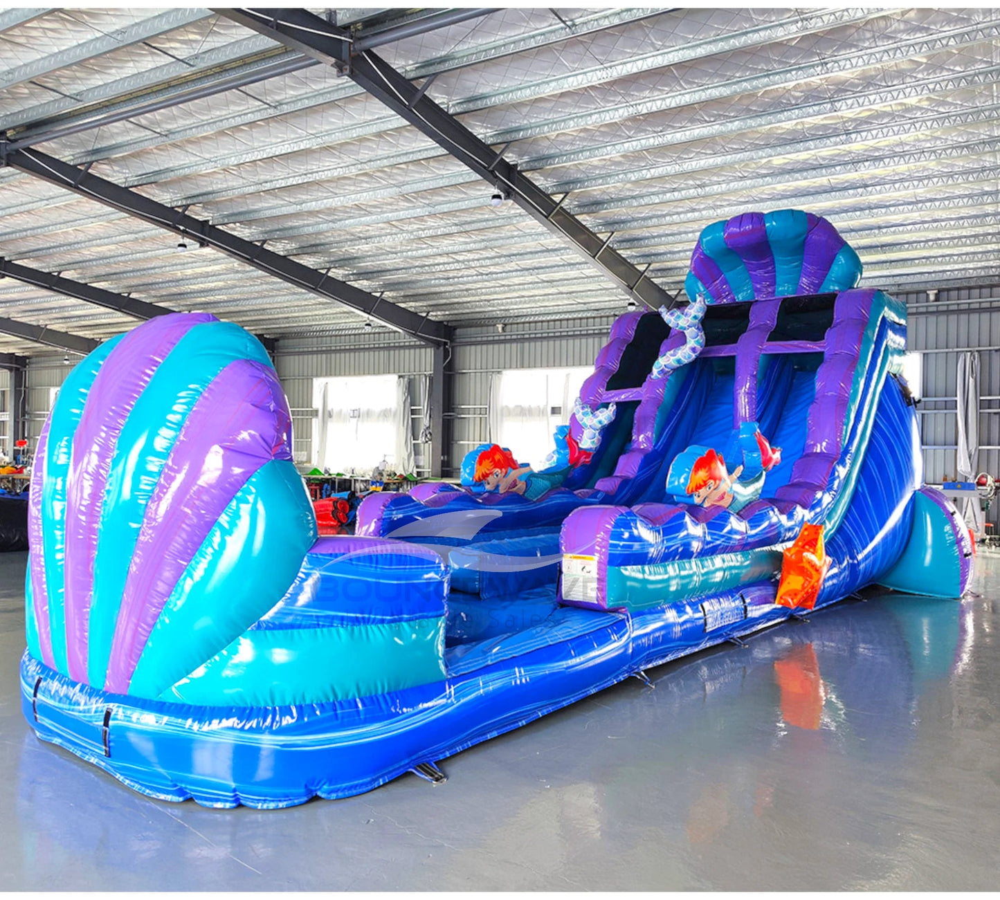 Large inflatable water slide with mermaid theme, featuring blue and purple colors, dual lanes, and shell-shaped top in indoor warehouse setting.