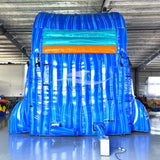 Rear view of Reef Racer inflatable water slide, displaying tall blue structure with shark silhouette and climbing area in warehouse environment.