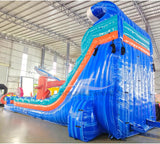 Large inflatable water slide with ocean theme, featuring blue sliding lanes, shark decorations, and a spacious splash pool area with octopus tentacles. The slide combines open and enclosed sections, set up in an indoor warehouse with metal roofing visible.