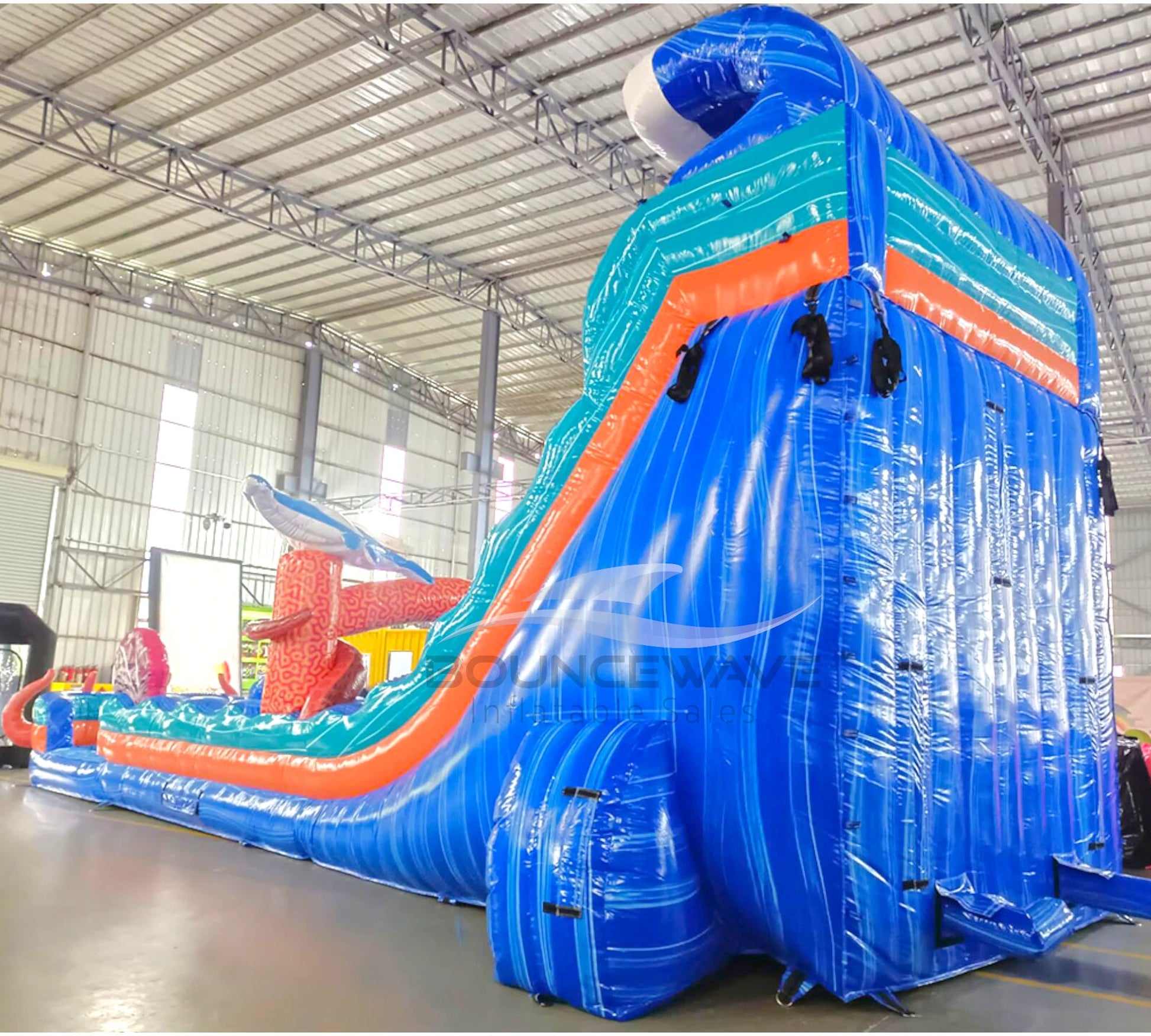 Large inflatable water slide with ocean theme, featuring blue sliding lanes, shark decorations, and a spacious splash pool area with octopus tentacles. The slide combines open and enclosed sections, set up in an indoor warehouse with metal roofing visible.