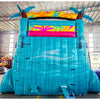 Large inflatable water slide with tropical theme, featuring turquoise sliding lanes, palm tree decorations, and a spacious splash pool area with colorful palm leaf designs. The slide combines open and enclosed sections, set up in an indoor warehouse with metal roofing and other equipment visible in the background.