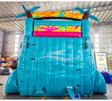 Large inflatable water slide with tropical theme, featuring turquoise sliding lanes, palm tree decorations, and a spacious splash pool area with colorful palm leaf designs. The slide combines open and enclosed sections, set up in an indoor warehouse with metal roofing and other equipment visible in the background.