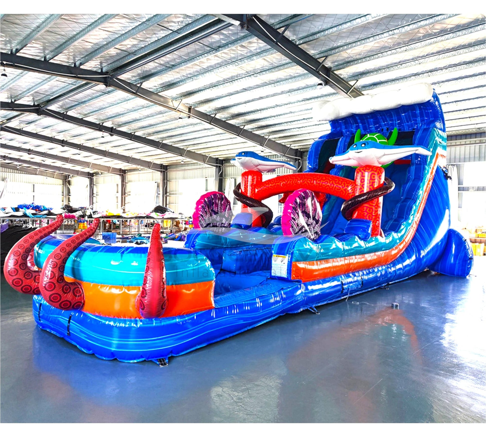 Large inflatable water slide with ocean theme, featuring vibrant blue colors, shark and stingray decorations, and a spacious splash pool area with octopus tentacles. The slide combines open and enclosed sections, set up in an indoor warehouse with metal roofing visible.