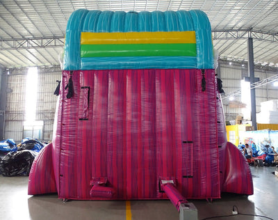 Rear view of 20ft Summer Luau Hybrid DL water slide featuring blue, green, and pink colors, with a multi-colored top section. The slide is set up in an indoor facility with metal roofing and other inflatable products visible in the background.