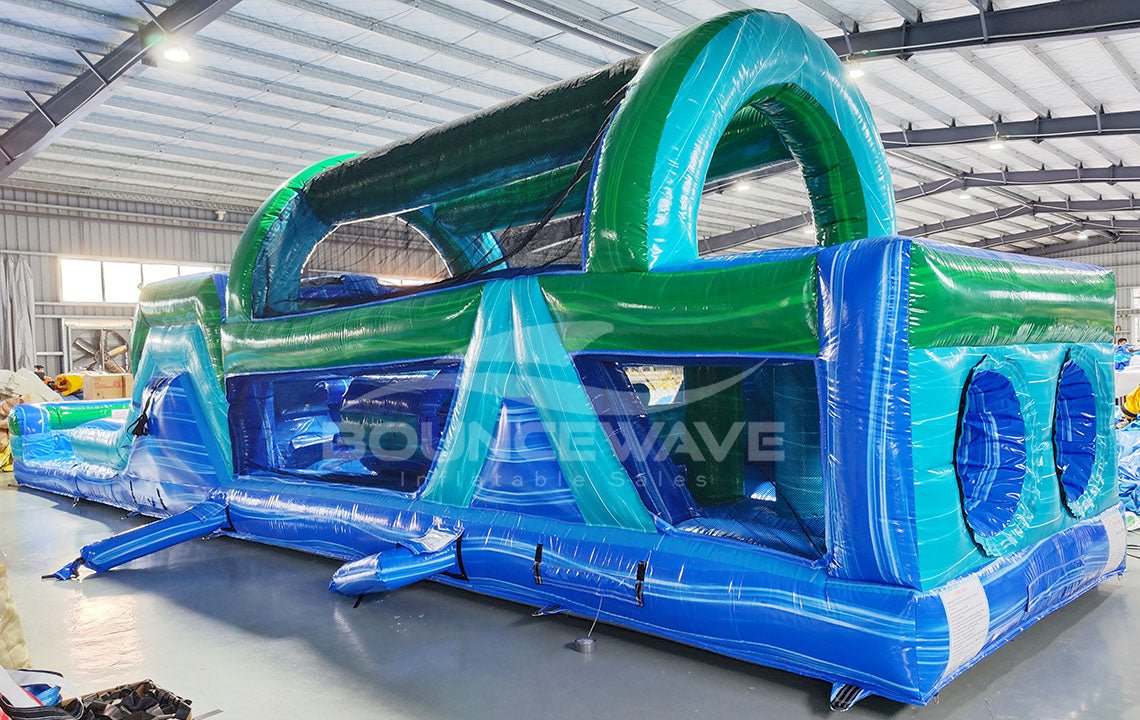 46ft Island Drop Hybrid Inflatable Obstacle Course side angle displaying long blue base, green accents, multiple challenges, and curved slide at the end
