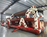 Wide-angle view of 46ft Rodeo Rush Commercial Hybrid Inflatable Obstacle Course in a large warehouse. The inflatable displays a cow-print design with a large cow figure arching over the structure, dual slides, and multiple obstacles. Various other inflatable products and warehouse equipment are visible, showcasing its size and commercial setting.