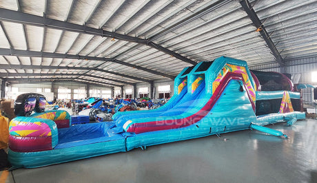 Wide-angle view of 53ft XL Island Flow Commercial Hybrid Inflatable Obstacle Course set up in a large indoor warehouse. The inflatable features dual blue slides, inflatable palm trees, and a long turquoise base with colorful accents. A large inflatable sunglasses with a tropical scene sits atop the structure. Various other inflatable products and storage containers are visible in the foreground and background.