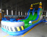 Front view of 53ft XL Radioactive Run Commercial Hybrid Inflatable Obstacle Course showing the entrance area. Features a large yellow and black striped arch, dual circular entrances with blue trim, and a diamond plate pattern base. The inflatable includes green slides and multiple obstacles. Set in a warehouse with concrete flooring and other inflatable products visible.