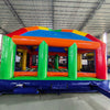 Wide-angle view of Mega Fun Dome Commercial Grade Bounce House in a spacious indoor facility. The inflatable features a rounded blue top with colorful panels, orange support beams, and green pillars. The entrance has a small orange archway. The image showcases the bounce house's size relative to the warehouse, with metal roofing, shelving units, and other inflatable products visible in the background.