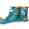Wide-angle view of Akua Falls Commercial Econo Inflatable Combo (Wet/Dry) in a spacious warehouse. Shows the full structure including the blue wave-shaped body with white patterns, brown palm tree pillars, yellow fronds, curved slide, and splash pool. Warehouse interior with metal roofing, shelving units, and other inflatable products visible, providing scale context.