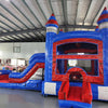 Front view of Baja Castle Econo Combo, showing bounce house entrance, slide, and splash pool. Features blue and red castle design with safety instructions visible. Set up in an indoor warehouse with other inflatables and equipment in the background.
