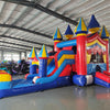 Wide-angle view of the Carnival Econo Combo featuring a colorful bounce house with multiple spires, attached slide, and large splash pool area. The inflatable is set up in a spacious indoor warehouse with metal roofing, bright lighting, and other inflatable products visible in the background.