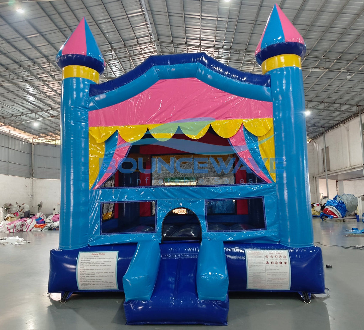 Front view of Carnival Pink Commercial Grade Bounce House set up in a large indoor warehouse. Features a vibrant blue castle-shaped structure with pink and yellow accents. Three turrets with blue and pink conical tops adorn the bounce house. The entrance has a yellow scalloped awning and mesh windows. A blue slide is attached to the side. The "BOUNCEWAVE" logo is visible on the front.