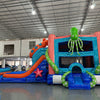 side view of Deep Sea Dive Dual Lane Commercial Inflatable Combo set up in a large indoor facility. Highlights the giant orange octopus, green tentacles, dual blue slides, and blue wave-patterned base with a splash pool. Safety instructions visible on the front. Warehouse interior with metal roofing and various equipment provides scale context.