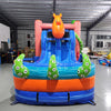 Front view of Deep Sea Dive Dual Lane Commercial Inflatable Combo (Wet/Dry) set up in a large indoor warehouse. Features a giant orange octopus on top, dual blue slides, green tentacles with eyes, and a blue wave-patterned base with a splash pool. Pink and orange coral-like patterns decorate the sides. Various inflatable products and warehouse equipment visible in the background.