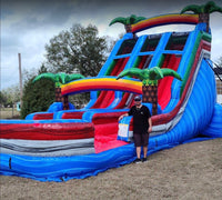 Bounce With It Inflatables testimonial: Eddie D. standing beside a large tropical-themed inflatable water slide with vibrant blue, pink, and green colors, showcasing impressive outdoor entertainment setup