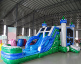 Large inflatable Euro Green Castle Dual Lane Commercial Combo featuring a castle-themed design with white towers, blue turrets, and green conical tops. The structure includes dual blue slides, a bounce house area with mesh windows, and a circular splash pool. Set up in a spacious warehouse with metal roofing and other inflatable products visible in the background.