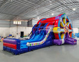 Wide-angle view of Ferris Wheel Dual Lane Commercial Inflatable Combo (Wet/Dry) in a spacious warehouse setting. Showcases the full structure including the Ferris wheel design, dual blue slides, and multicolored base. The warehouse interior is visible with metal roofing, walls, and other inflatable products in the background, providing scale and context for the combo's size.
