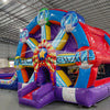 Large colorful inflatable combo featuring a Ferris wheel design with dual slides. The structure has vibrant blue, red, purple, and yellow colors. Central Ferris wheel element has multicolored spokes and a star-shaped center. Two blue slides flank the wheel, leading to a splash pool area. Set up in a spacious indoor warehouse with metal roofing visible.