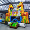 Wide-angle view of the Giraffe Safari Econo Combo in an indoor warehouse. Features two large giraffe inflatables, a bounce house with animal artwork, blue slide, and green obstacles. Other inflatable products and warehouse equipment visible in the background.