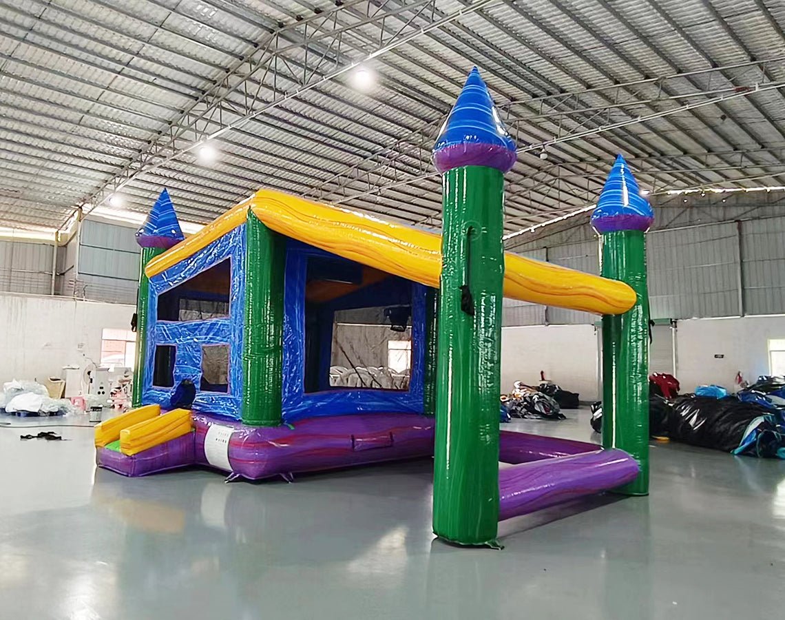 Front view of Goombay Commercial Grade Canopy Bounce House featuring a vibrant blue inflatable structure with four tall pillars topped by white cylindrical turrets and blue conical roofs. The bounce area has yellow walls with mesh windows. A yellow canopy extends between the turrets, creating a covered play space. Set up in a large indoor warehouse with metal roofing and other inflatable products visible in the background.