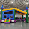 Three-quarter view of Goombay Commercial Grade Canopy Bounce House highlighting its blue inflatable frame, yellow bounce area walls, and yellow canopy top. Four tall blue pillars with white cylindrical turrets and blue conical roofs anchor the corners. Mesh windows allow visibility into the play area. The bounce house is displayed in a large indoor facility with metal roofing, fluorescent lighting, and other inflatable products in the background.