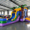 Wide-angle view of Goombay Splash Econo Combo showing bright blue, yellow, and purple design with inflatable palm trees, curved slide, and large splash pool area. The combo is set up in a spacious indoor warehouse with metal roofing, bright lighting, and other inflatable products visible in the background.
