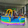 Front angled view of Goombay Curved Dual Lane Commercial Inflatable Combo (Wet/Dry) in a spacious warehouse. Highlights the yellow climbing structure with blue accents, dual blue curved slides, inflatable palm trees, and circular multicolored splash pool. Warehouse interior visible with metal roofing, shelving units, and other inflatable products, providing scale and context for the combo's size.