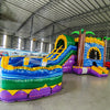 Wide-angle view of Goombay Curved Dual Lane Commercial Inflatable Combo (Wet/Dry) set up in a large indoor warehouse. Features dual blue curved slides, inflatable palm trees, and a tall yellow climbing structure. The large circular splash pool with multicolored sections is prominent in the foreground. Metal roofing, disco balls, and other warehouse equipment visible, providing context for its commercial use and size.