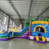 Side view of Goombay Curved Dual Lane Commercial Inflatable Combo (Wet/Dry) in a warehouse setting. Showcases the full structure including dual blue curved slides, yellow climbing wall with blue accents, inflatable palm trees, and circular multicolored splash pool. Other inflatable products and warehouse equipment visible in the background, demonstrating its scale in a commercial environment.