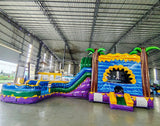 Side view of Goombay Curved Dual Lane Commercial Inflatable Combo (Wet/Dry) in a warehouse setting. Showcases the full structure including dual blue curved slides, yellow climbing wall with blue accents, inflatable palm trees, and circular multicolored splash pool. Other inflatable products and warehouse equipment visible in the background, demonstrating its scale in a commercial environment.